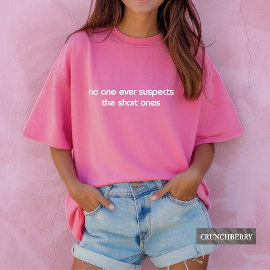 A woman wearing a bright pink oversized t-shirt with the white text 'no one ever suspects the short ones' on the front, paired with light blue denim shorts, standing against a textured pink wall. A 'CRUNCHBERRY' brand label is visible in the bottom right corner.