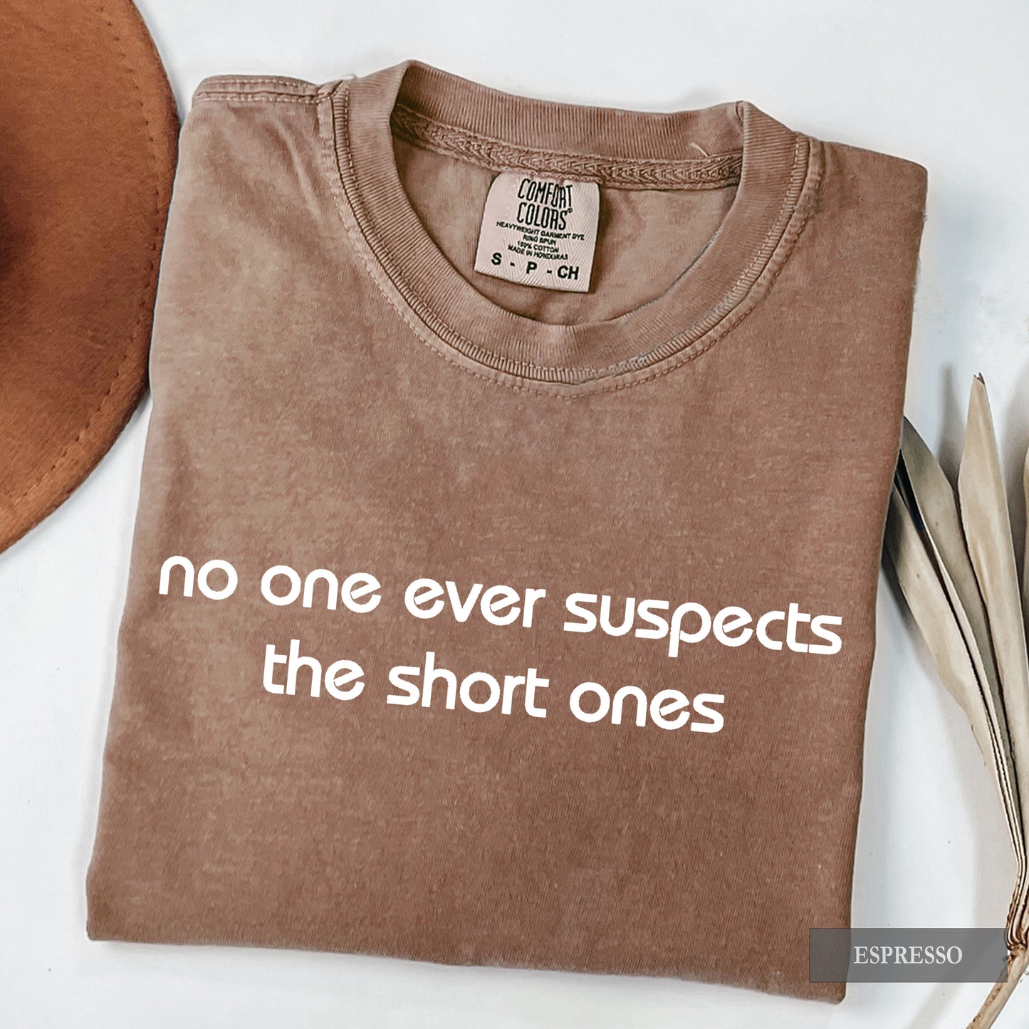 A folded brown t-shirt with white text reading 'no one ever suspects the short ones' on a white background, styled with a brown hat and dried leaves. The brand label reads 'Comfort Colors'.