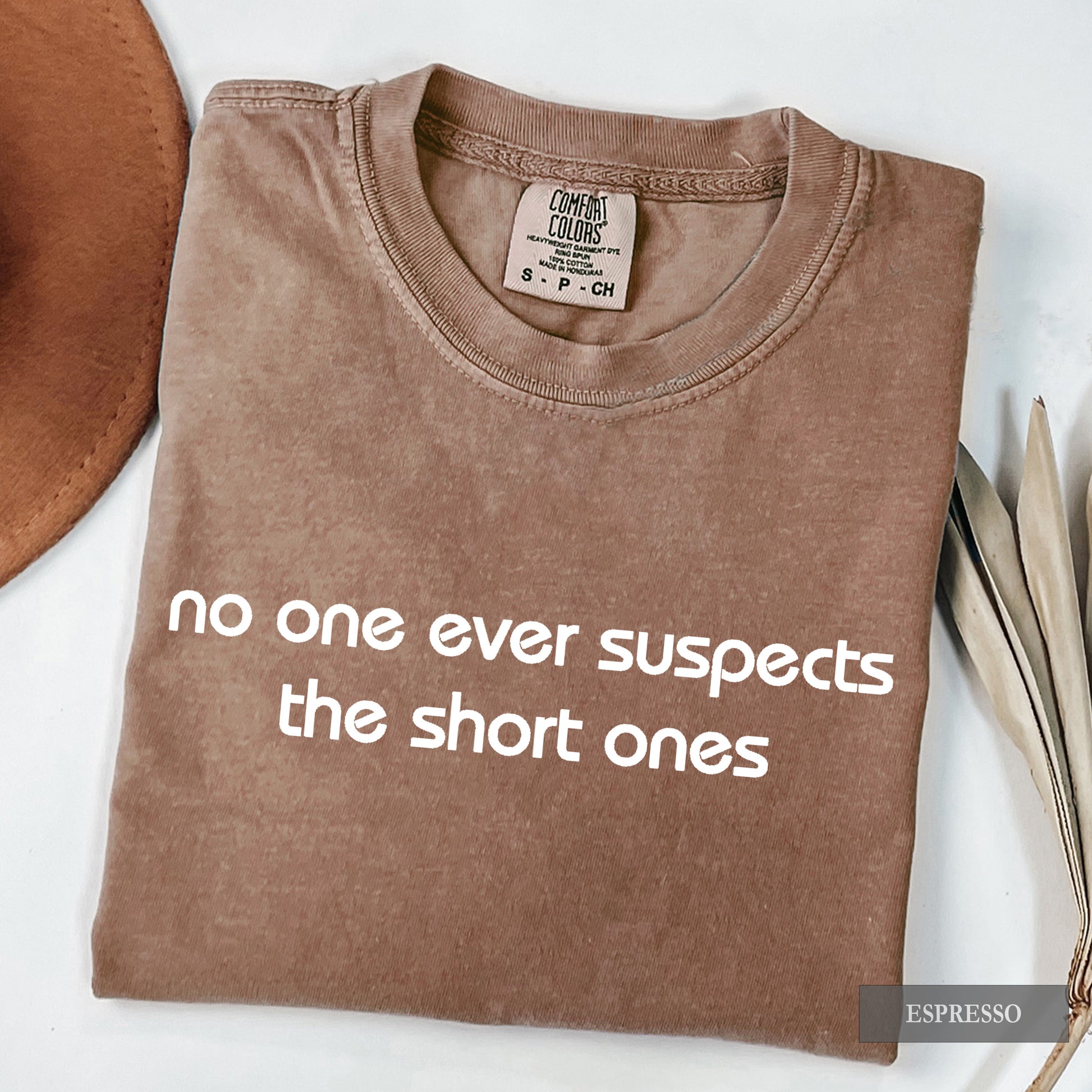 A folded brown t-shirt with white text reading 'no one ever suspects the short ones' on a white background, styled with a brown hat and dried leaves. The brand label reads 'Comfort Colors'.