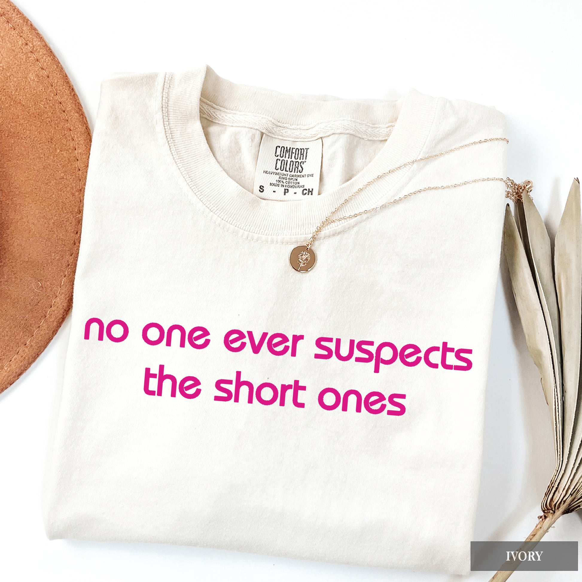 A folded ivory t-shirt with pink text reading 'no one ever suspects the short ones' on the front, styled with a gold pendant necklace, a brown leather circle, and dried leaves on a white background.