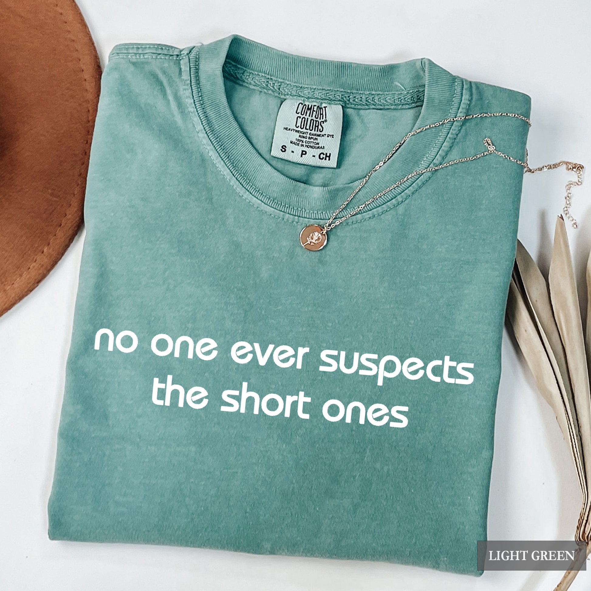 A folded light green t-shirt with the phrase 'no one ever suspects the short ones' printed in white across the front. The shirt is styled with a delicate silver necklace and a brown leather accessory on a white background.