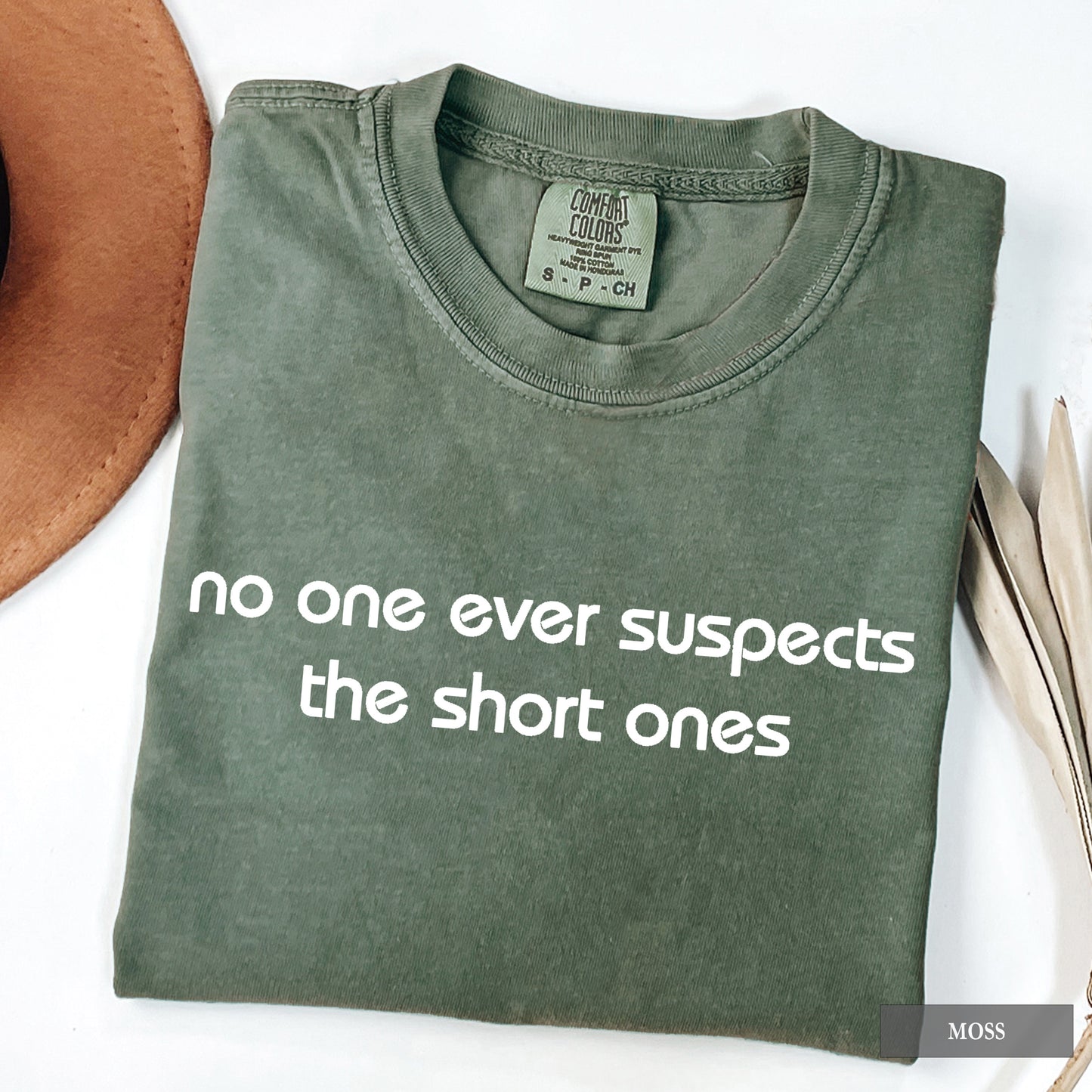 An olive green folded t-shirt with the phrase 'no one ever suspects the short ones' printed in white across the front. The t-shirt is displayed on a white surface with a brown hat partially visible on the left and dried leaves on the right.