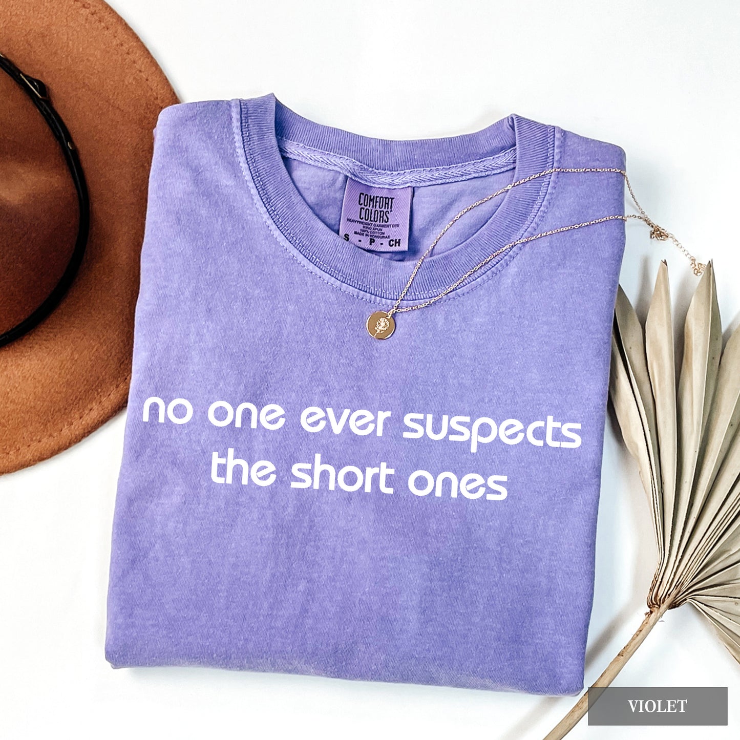 A folded lavender-purple t-shirt with white text reading 'no one ever suspects the short ones' is styled in a flat lay on a white background with a brown hat, sunglasses, a necklace, and dried palm leaves. A 'VIOLET' brand label is visible inside the collar.