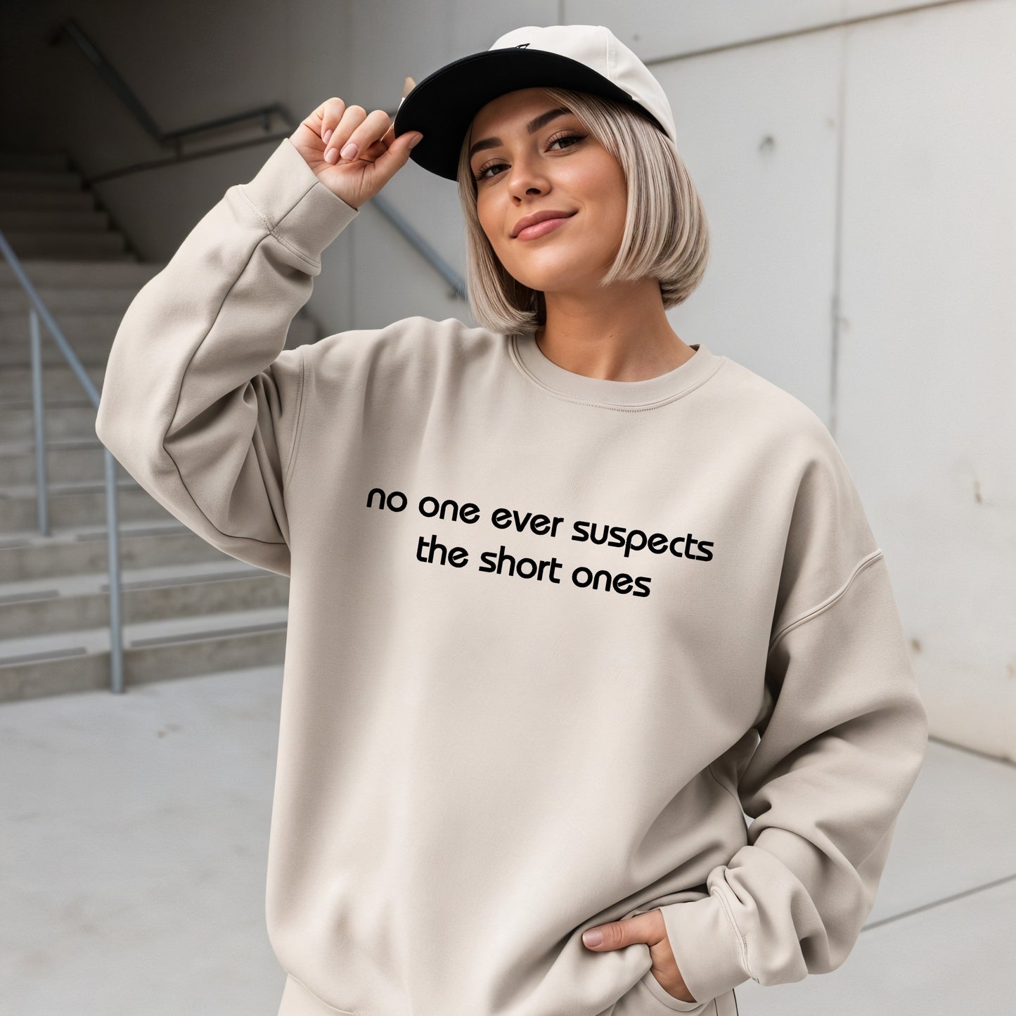 A woman wearing a beige crewneck sweatshirt with the black printed text 'no one ever suspects the short ones', styled with a black and white baseball cap, standing in an outdoor concrete setting.
