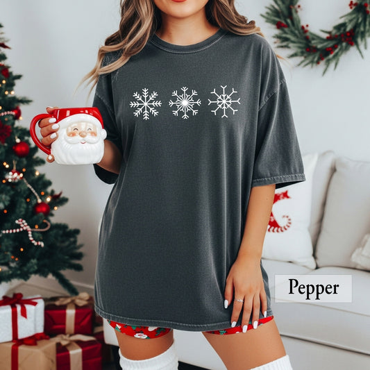 A woman wearing a pepper comfort colors t-shirt with three snowflakes on the front.