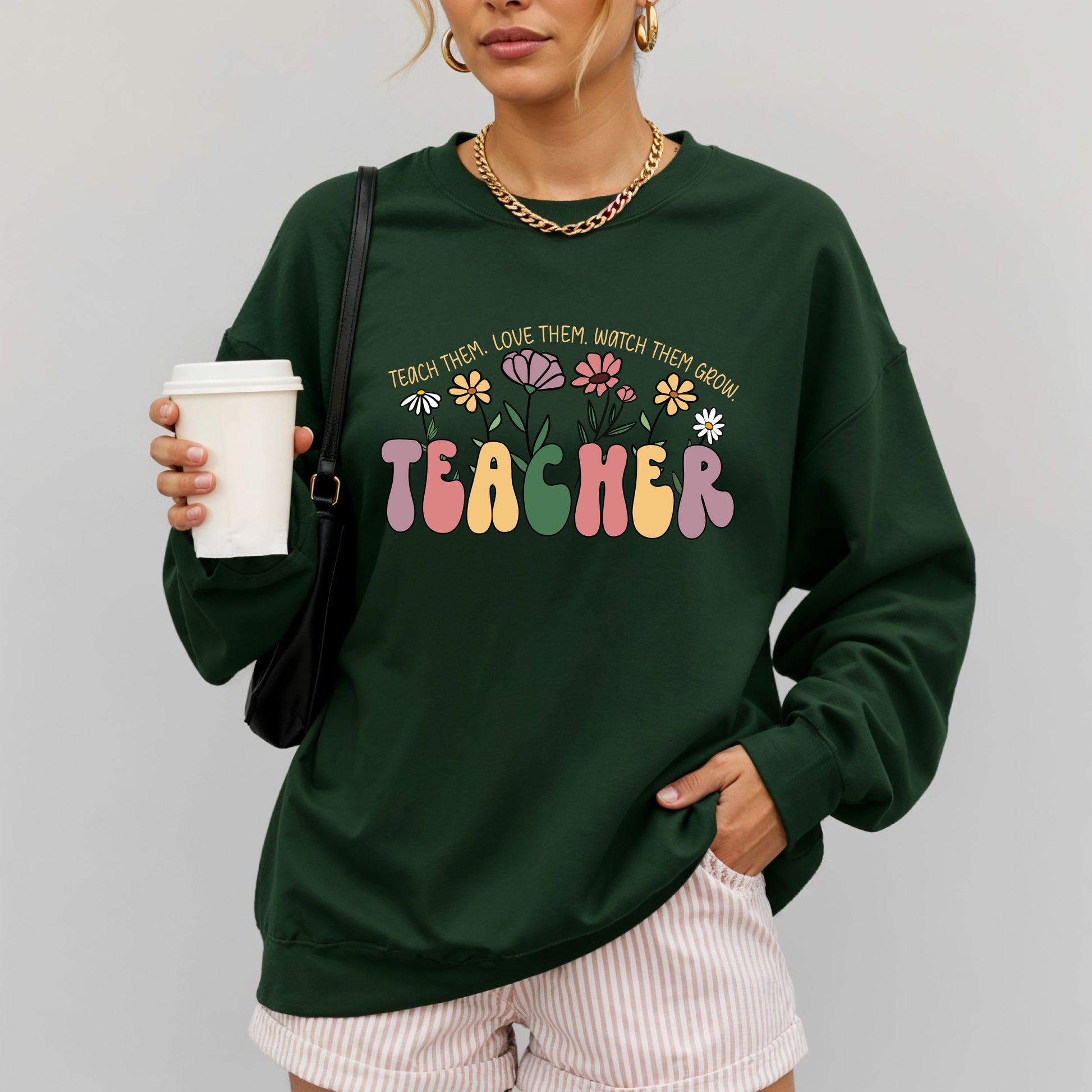 Person wearing a green sweatshirt with 'TEACHER' text and flowers, holding a coffee cup.