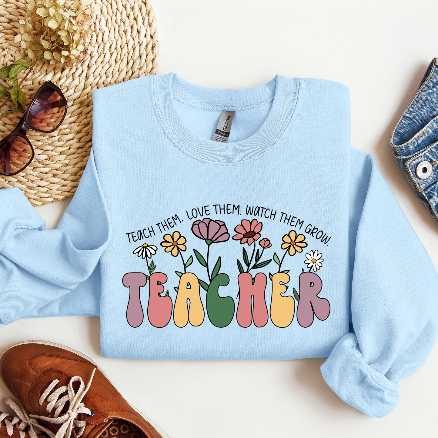 Light blue sweatshirt with colorful 'TEACHER' text and floral design on a white surface with accessories.