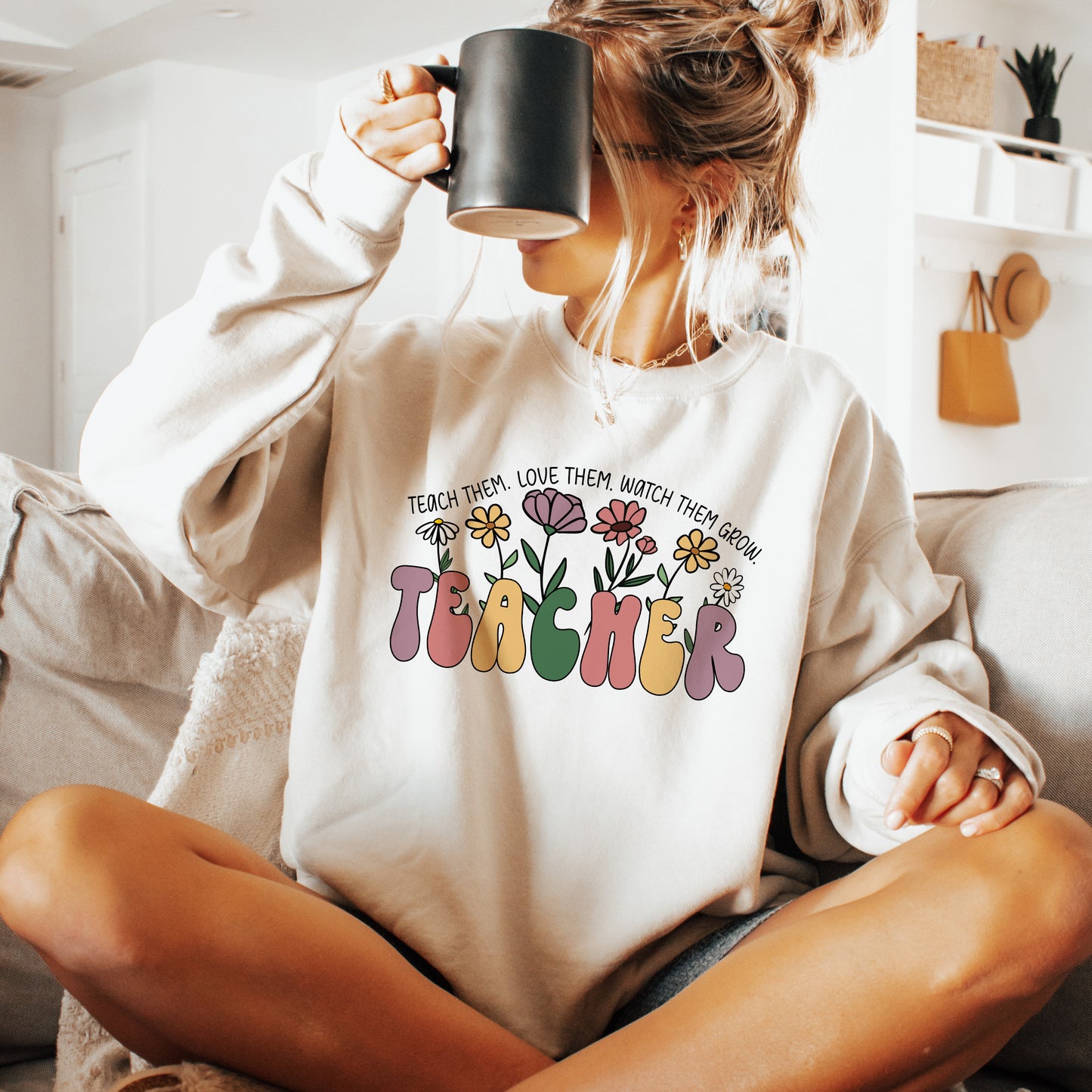 Person wearing a white sweatshirt with colorful 'TEACHER' design and floral elements, sitting on a couch.
