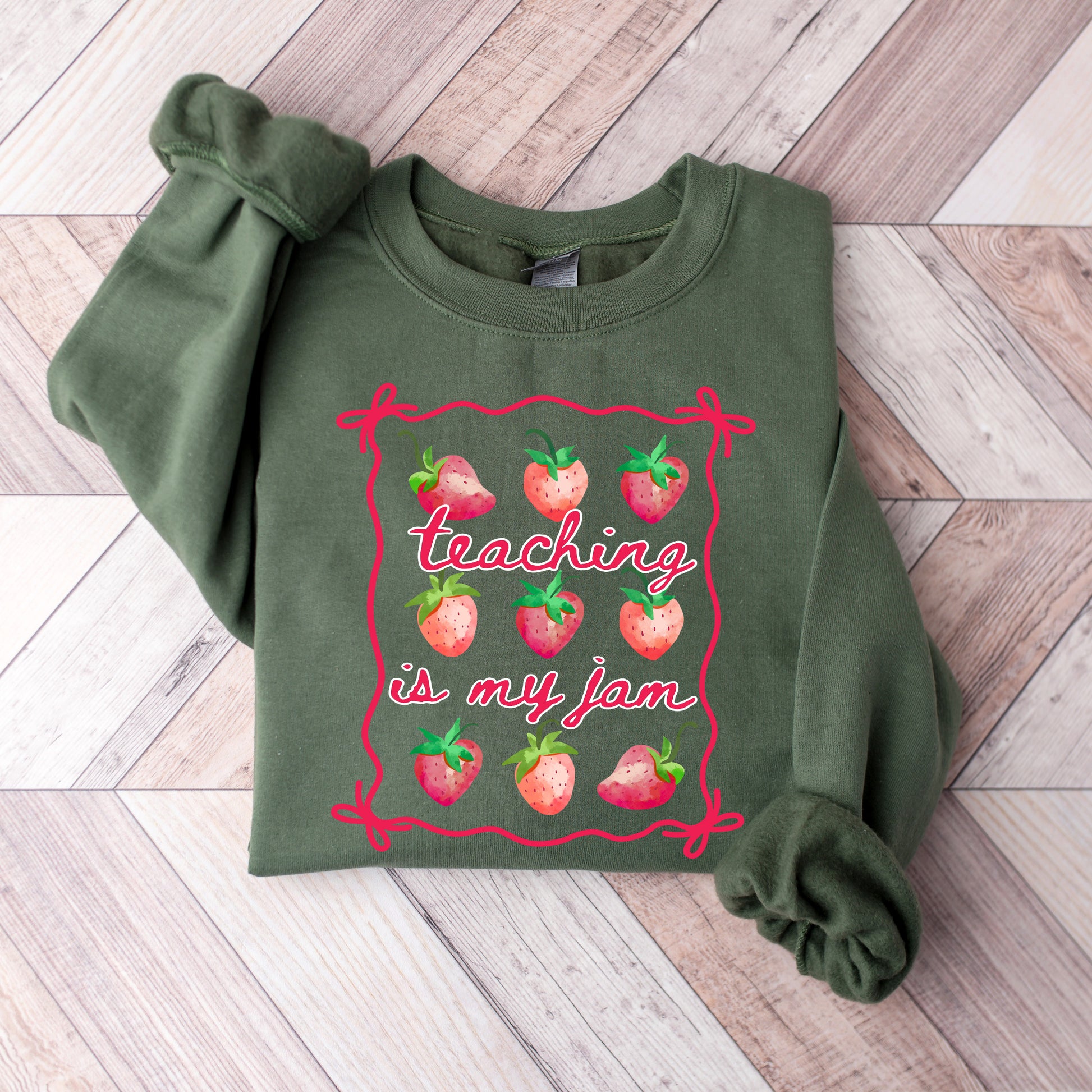 Green sweatshirt with 'teaching is my jam' text and strawberry graphics on a wooden surface