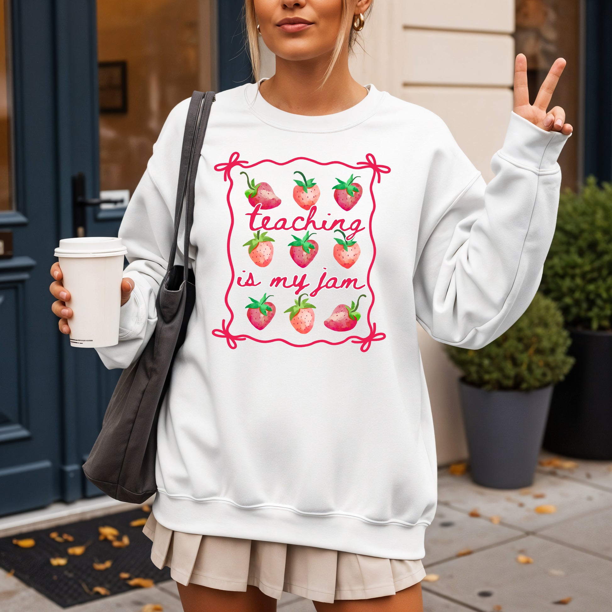 Person wearing a white sweatshirt with 'Teaching is my jam' design, holding a coffee cup outdoors.