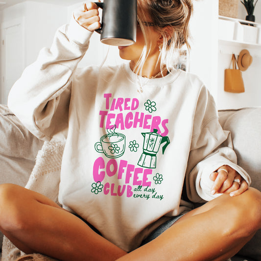 Person wearing a sweatshirt with 'Tired Teachers Coffee Club' text and graphics, holding a mug.