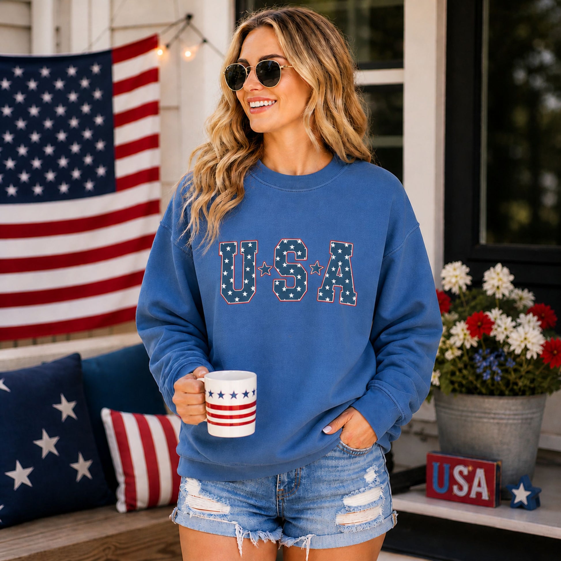Woman wearing a blue 'USA' sweatshirt holding a mug, standing in front of an American flag and flowers.