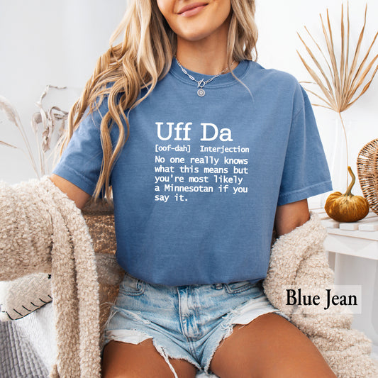 A woman wearing a blue graphic t-shirt with a white text definition print that reads 'Uff Da [oof-da] Interjection No one really knows what this means but you're most likely a Minnesotan if you say it.' She is sitting in a cozy indoor setting with a beige knit blanket draped over her shoulders and denim shorts.