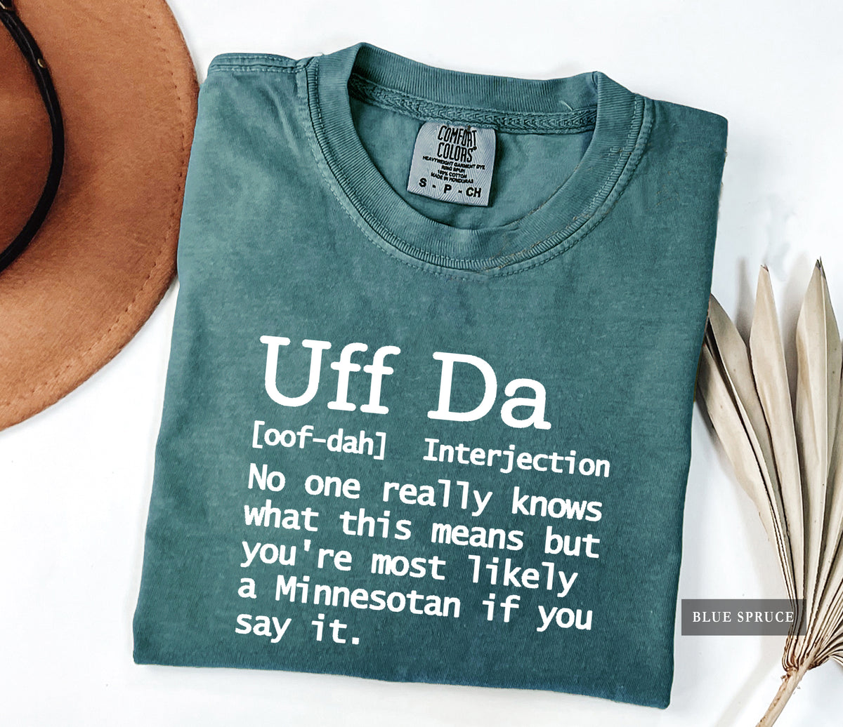 A folded slate blue-green t-shirt with white printed text reading 'Uff Da [oof-dah] Interjection No one really knows what this means but you're most likely a Minnesotan if you say it.' styled on a white background with a brown hat and dried palm leaves.