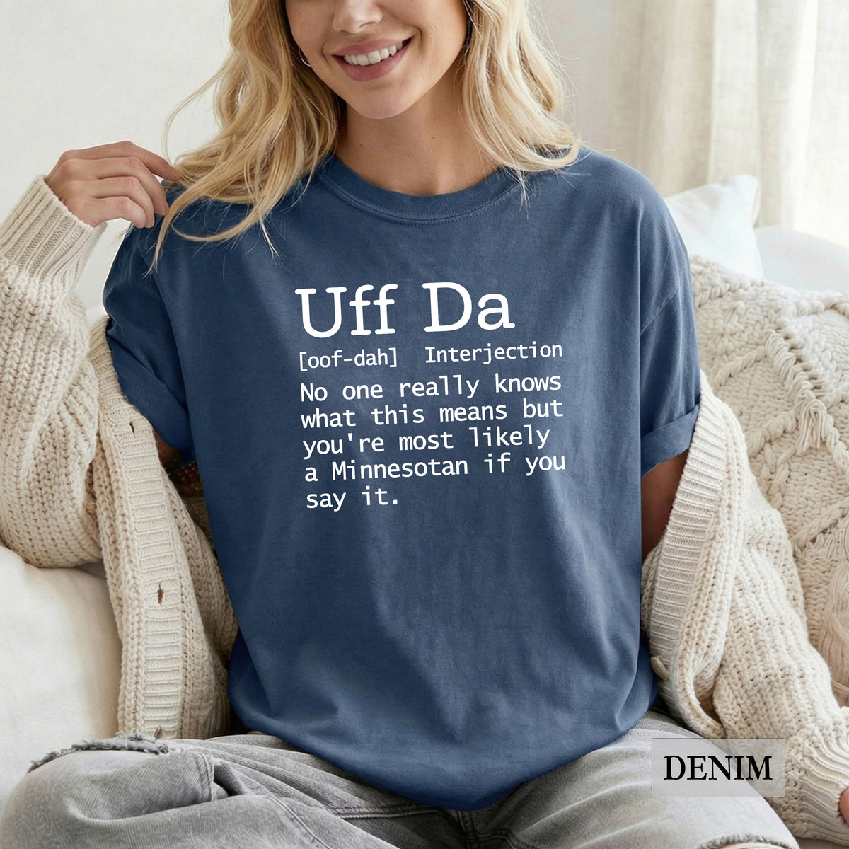 A woman wearing a navy blue t-shirt with a white printed definition of the word 'Uff Da' sits on a couch wrapped in a cream knit cardigan. The t-shirt features a white text graphic with a humorous definition of 'Uff Da' and the word 'DENIM' is printed on the bottom right.