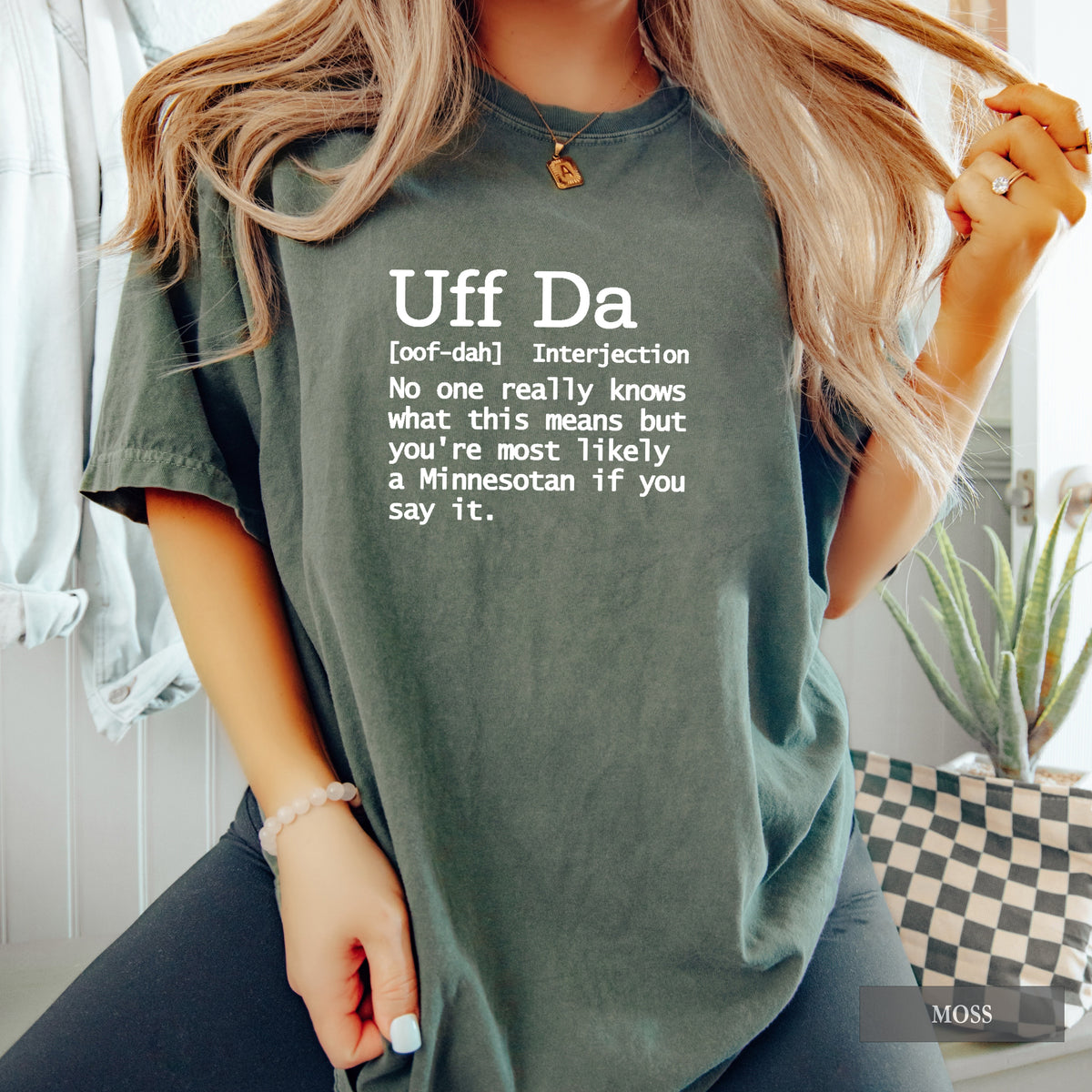 A woman wearing an oversized moss green graphic t-shirt with a white printed dictionary definition of 'Uff Da'. The background shows a cozy indoor setting with a checkered plant pot and light-colored walls.