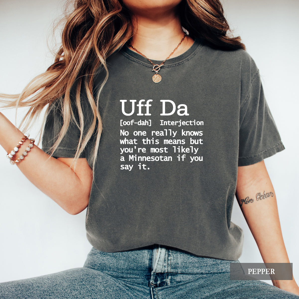A woman wearing a dark charcoal gray graphic t-shirt with white printed text defining the slang phrase 'Uff Da'. The t-shirt is labeled as 'PEPPER' in a gray overlay at the bottom right.