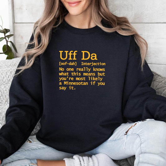 A woman wearing a black crewneck sweatshirt with a yellow printed dictionary-style definition that reads 'Uff Da [cof-dah] Interjection No one really knows what this means but you're most likely a Minnesotan if you say it.' She is sitting on a gray couch wearing light-wash ripped jeans.