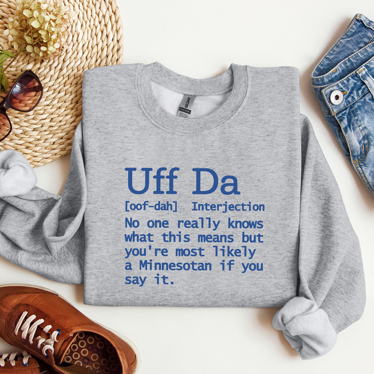 A heather gray crewneck sweatshirt with blue printed text defining 'Uff Da' is shown in a flat-lay arrangement with a straw hat, sunglasses, brown shoes, and blue jeans on a white background.