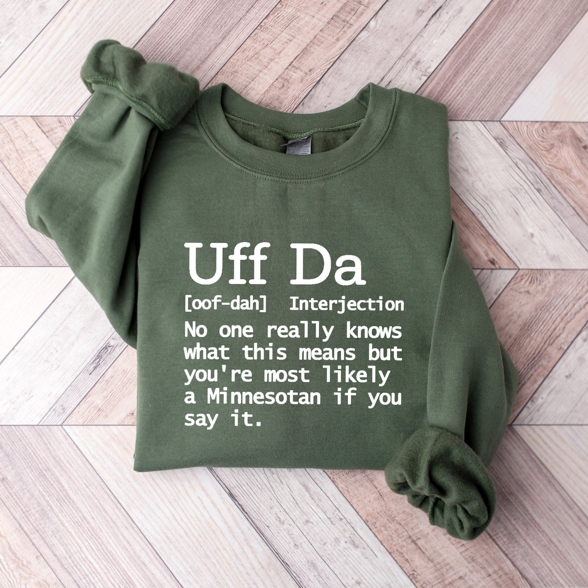 An olive green long-sleeve sweatshirt with white printed text defining 'Uff Da', laid flat on a light wood plank background.