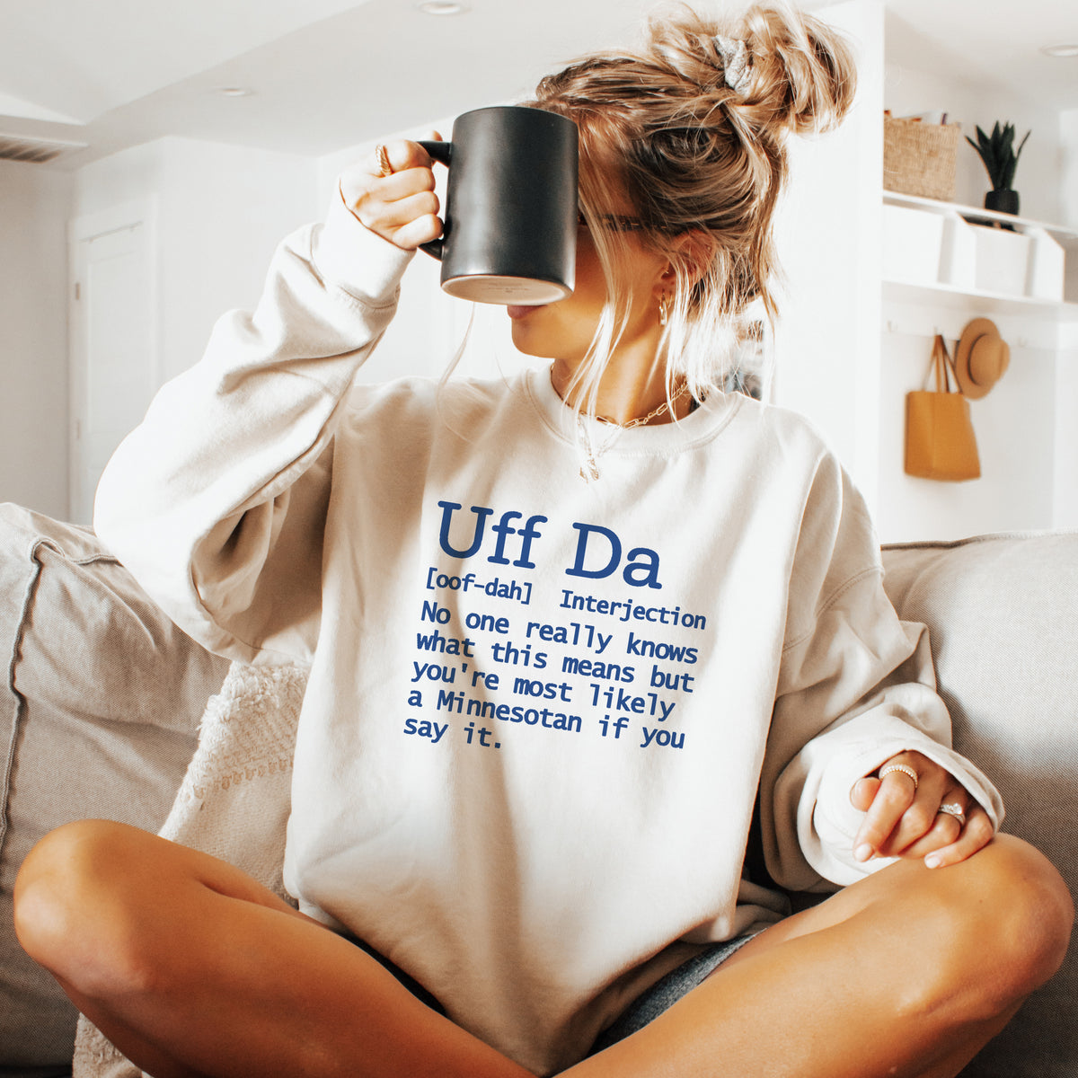 A woman sitting cross-legged on a couch in a bright living room, wearing a cream sweatshirt with a blue dictionary-style definition print that reads 'Uff Da' and holding a black mug.