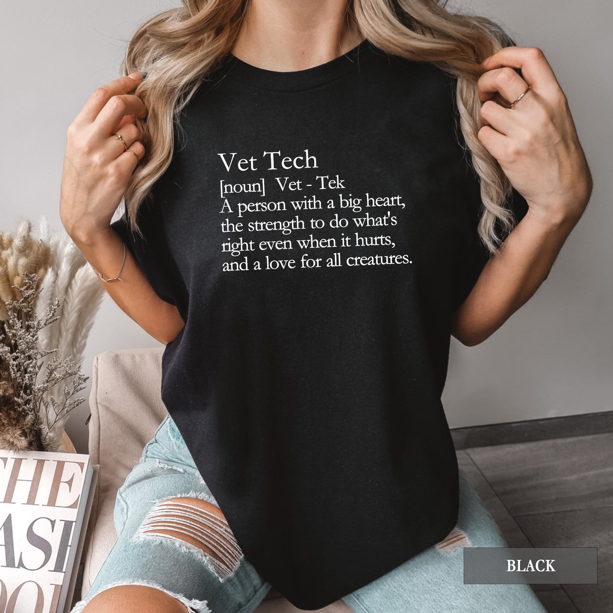 A woman wearing a black graphic t-shirt with a white printed definition of 'Vet Tech' on the chest, paired with light-wash distressed denim shorts in a cozy indoor setting.
