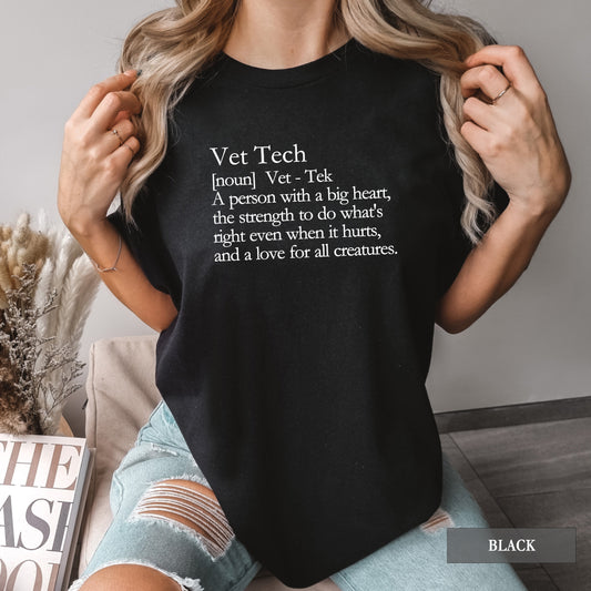 A woman wearing a black graphic t-shirt with a white printed definition of 'Vet Tech' on the chest, paired with light-wash distressed denim shorts in a cozy indoor setting.