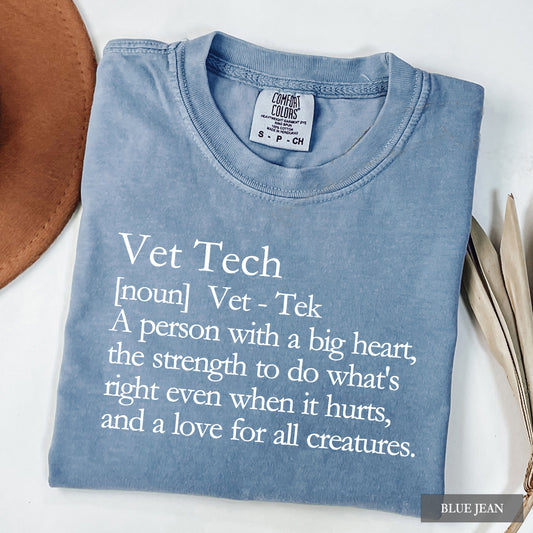 A folded light blue t-shirt with white printed text defining 'Vet Tech' humorously, styled on a white background with a brown hat and dried leaves.