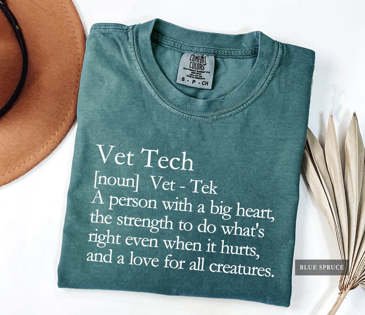 A folded slate blue t-shirt with white printed text defining 'Vet Tech' is displayed on a white background with a brown hat and dried palm leaves as props. The brand 'Comfort Colors' is visible on the inner neck label.