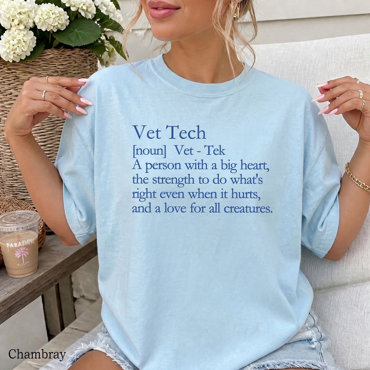 A woman wearing a light chambray blue graphic t-shirt with a blue text print that reads 'Vet Tech [noun] Vet - Tek A person with a big heart, the strength to do what's right even when it hurts, and a love for all creatures.' She is sitting on a white couch with a coffee cup and flowers in the background.