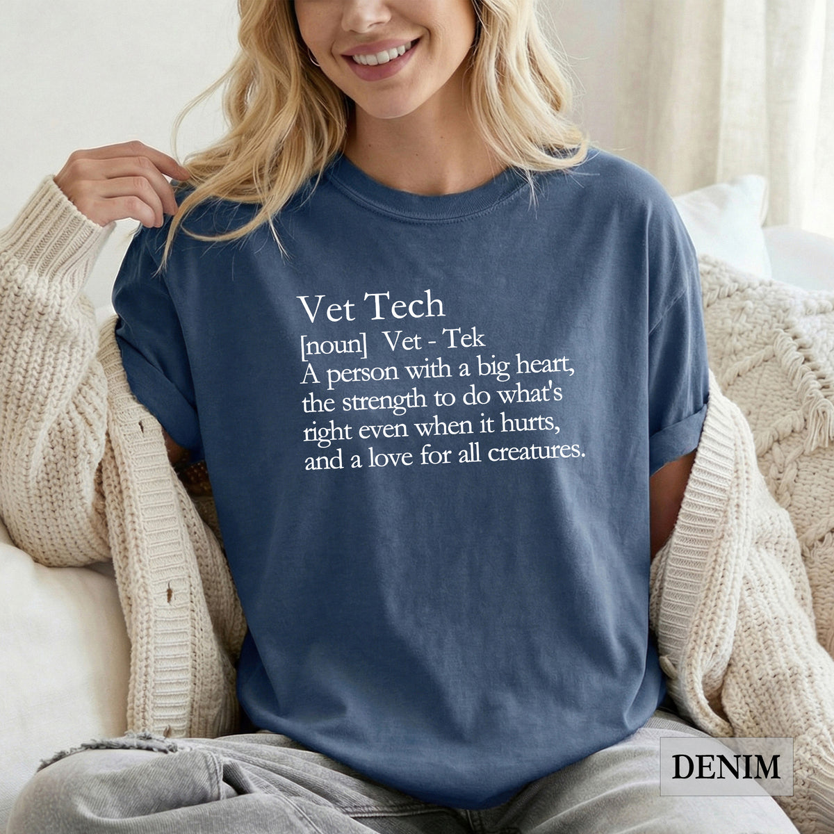 A woman wearing a navy blue graphic t-shirt with a white printed definition of 'Vet Tech' sitting on a couch with a cream knit cardigan.