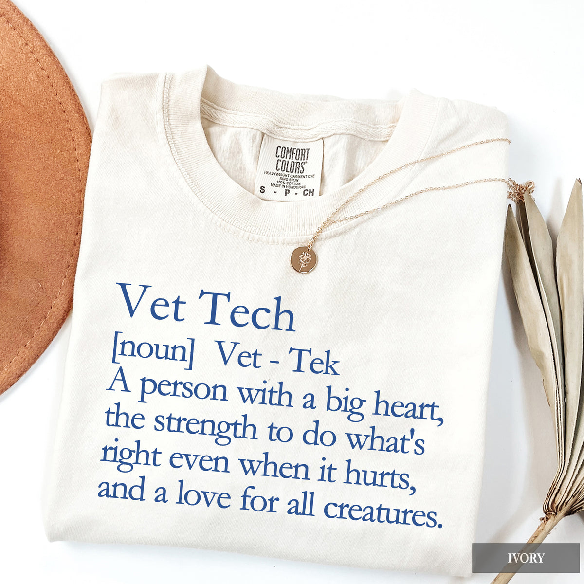 An ivory folded t-shirt with blue text reading 'Vet Tech' and a dictionary definition, styled with a gold coin necklace and dried palm leaves on a white background.