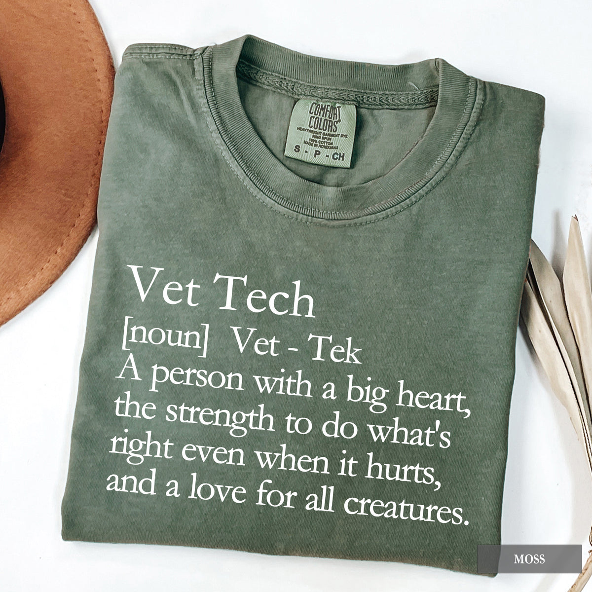 An olive green folded t-shirt with a white printed definition of 'Vet Tech' on the front, styled on a white background with a brown hat and dried leaves as props.
