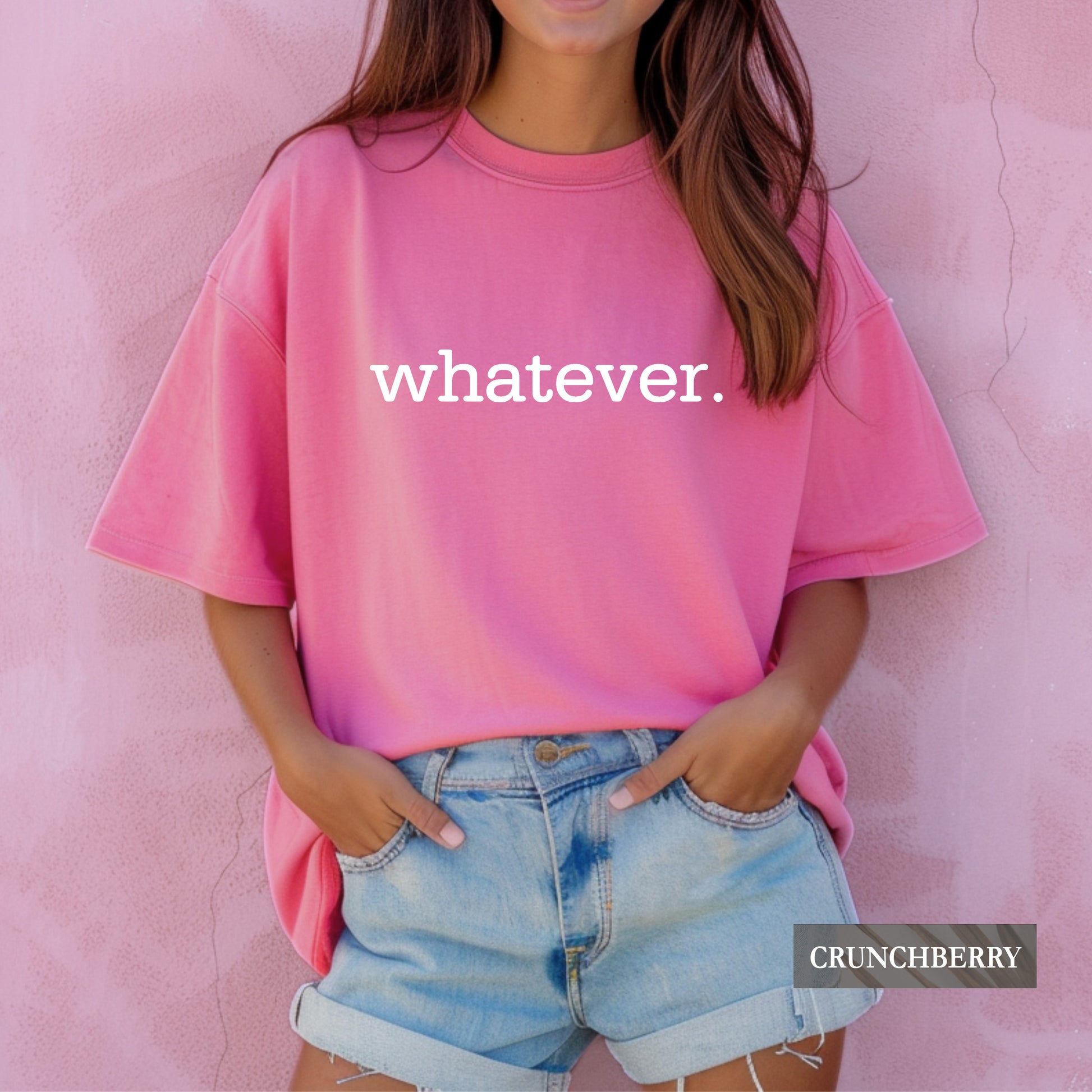 A woman wearing a bright pink oversized t-shirt with the white text 'whatever' on the front, paired with light blue denim shorts, standing against a textured pink wall. A 'CRUNCHBERRY' brand label is visible in the bottom right corner.