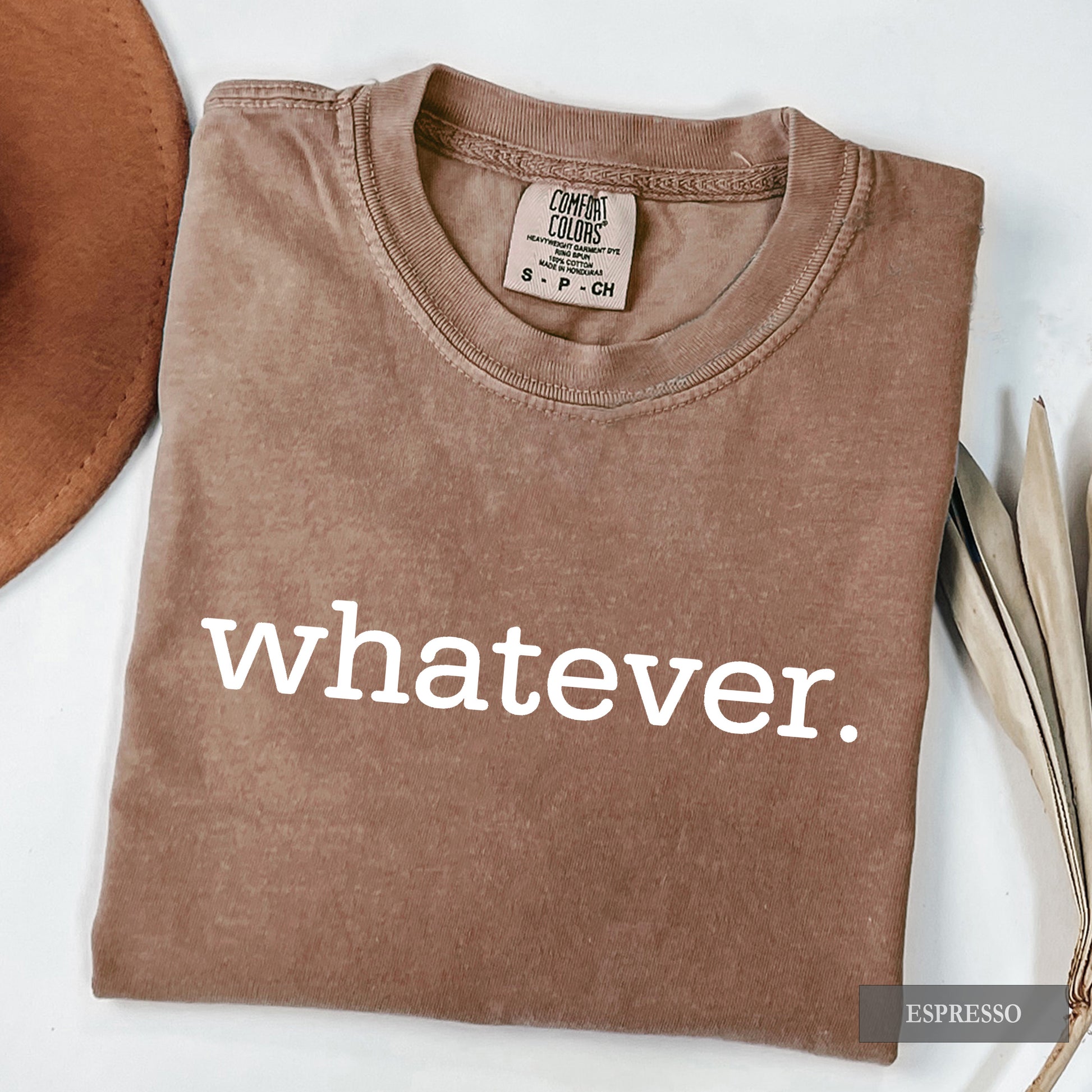 A folded brown t-shirt with the word 'whatever.' printed in white lowercase letters across the front, displayed on a white background with decorative props.