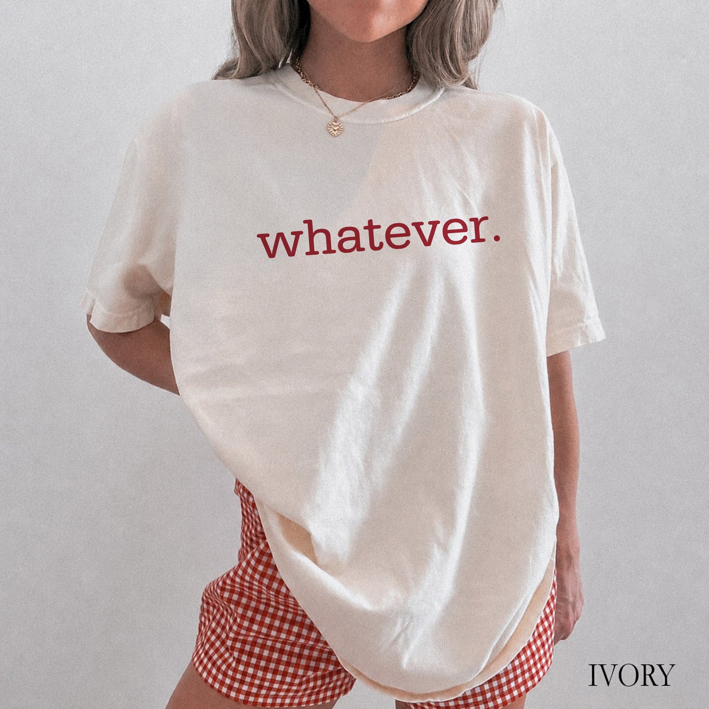 A person wearing an oversized ivory T-shirt with the word "whatever." printed across the chest in red lowercase letters, paired with red-and-white gingham shorts.