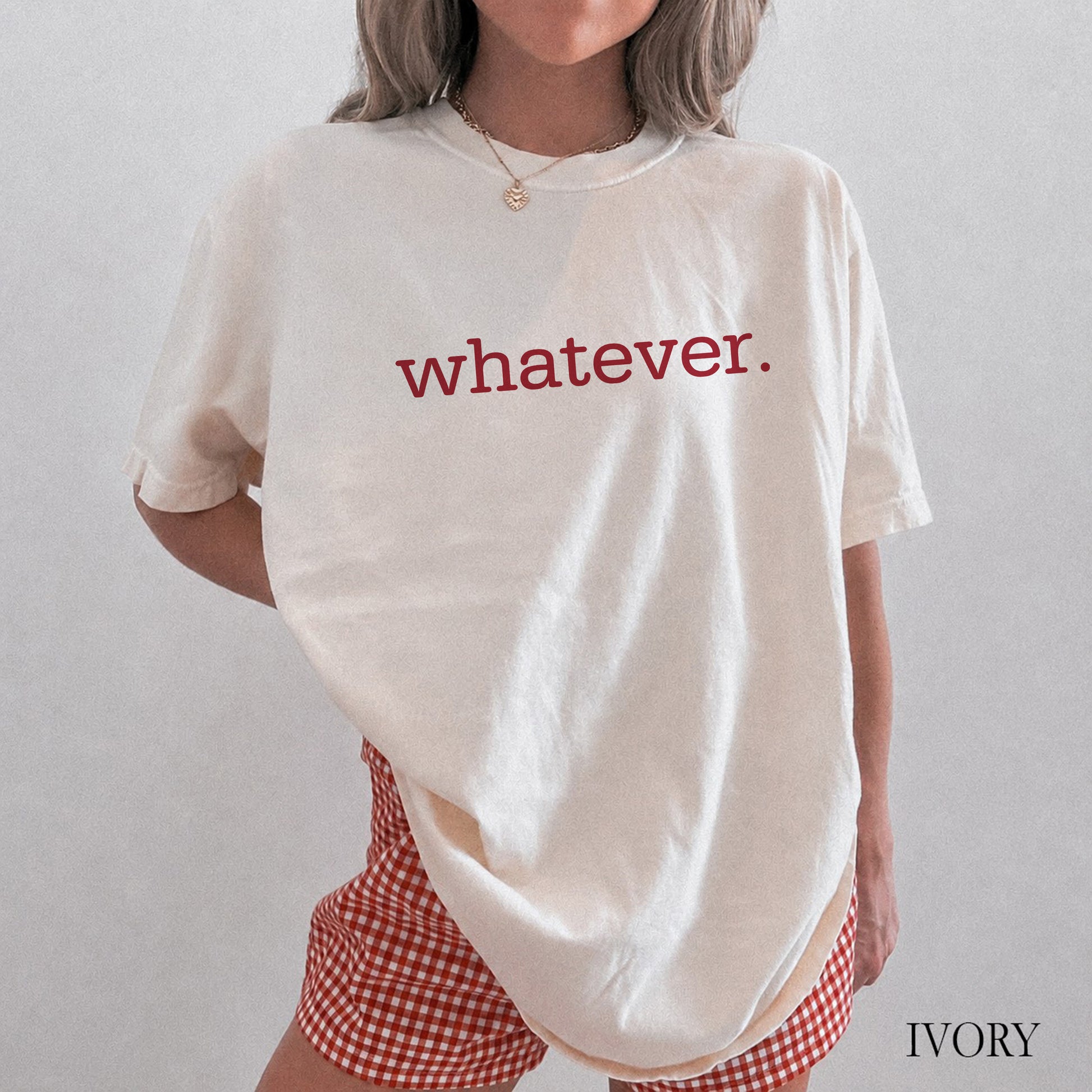 A person wearing an oversized ivory T-shirt with the word "whatever." printed across the chest in red lowercase letters, paired with red-and-white gingham shorts.
