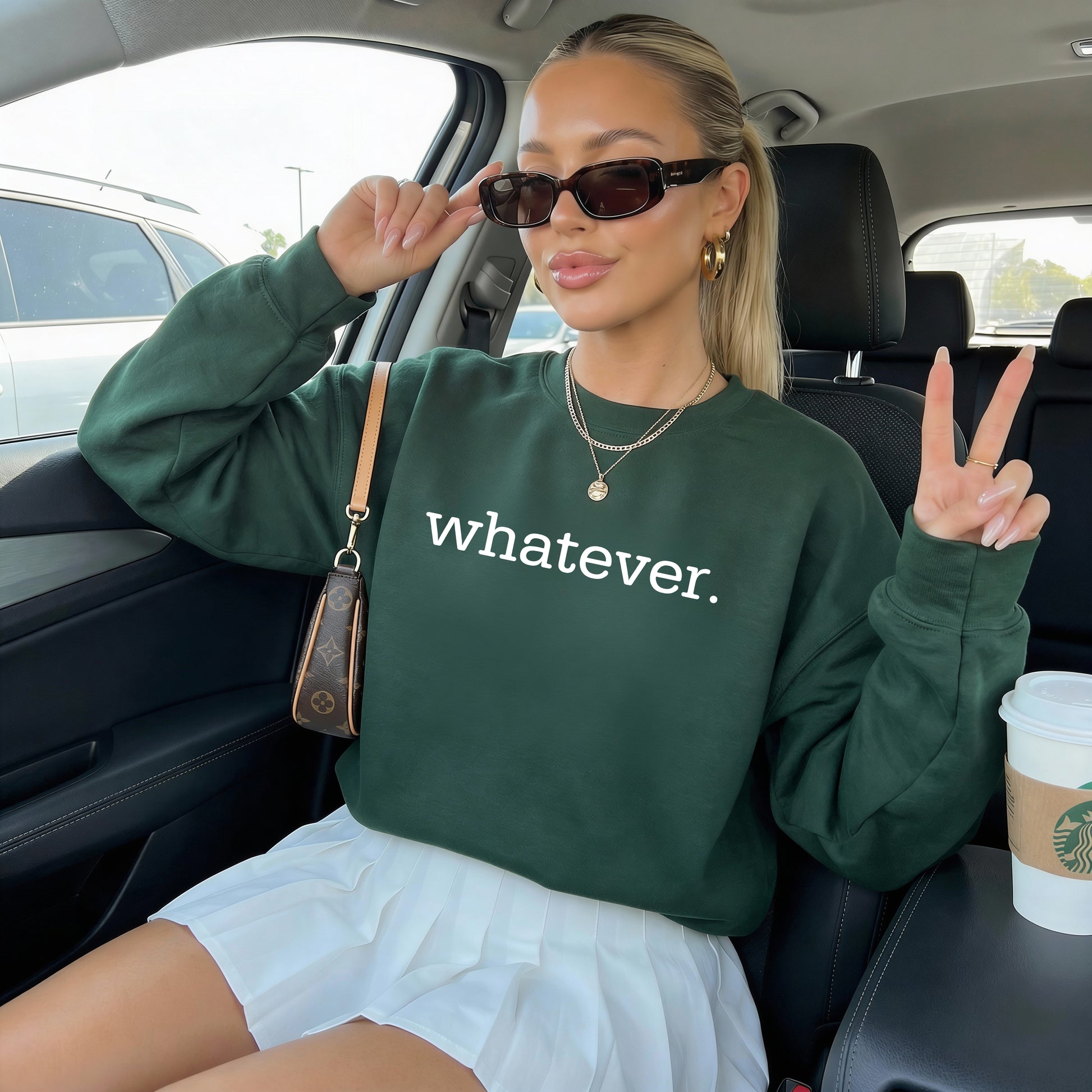 A woman sitting in a car wearing a dark green crewneck sweatshirt with the word 'whatever.' printed in white across the chest, paired with a white skirt and sunglasses.