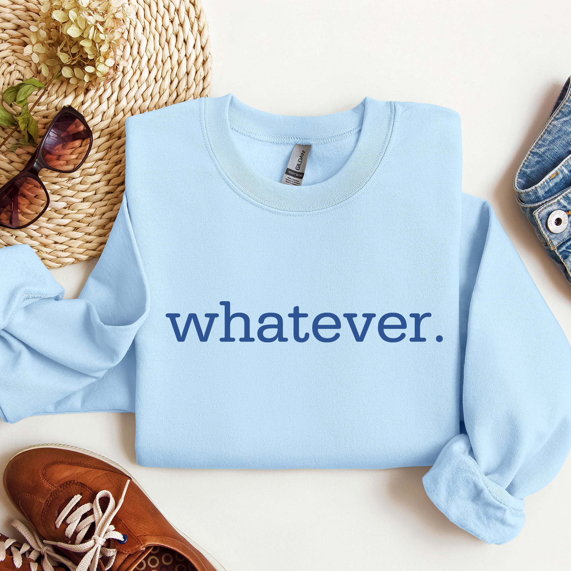 A light blue crewneck sweatshirt with the word 'whatever.' printed in dark blue across the chest, styled in a flat lay with sunglasses, a woven hat, a denim shoe, and a brown leather shoe on a white background.