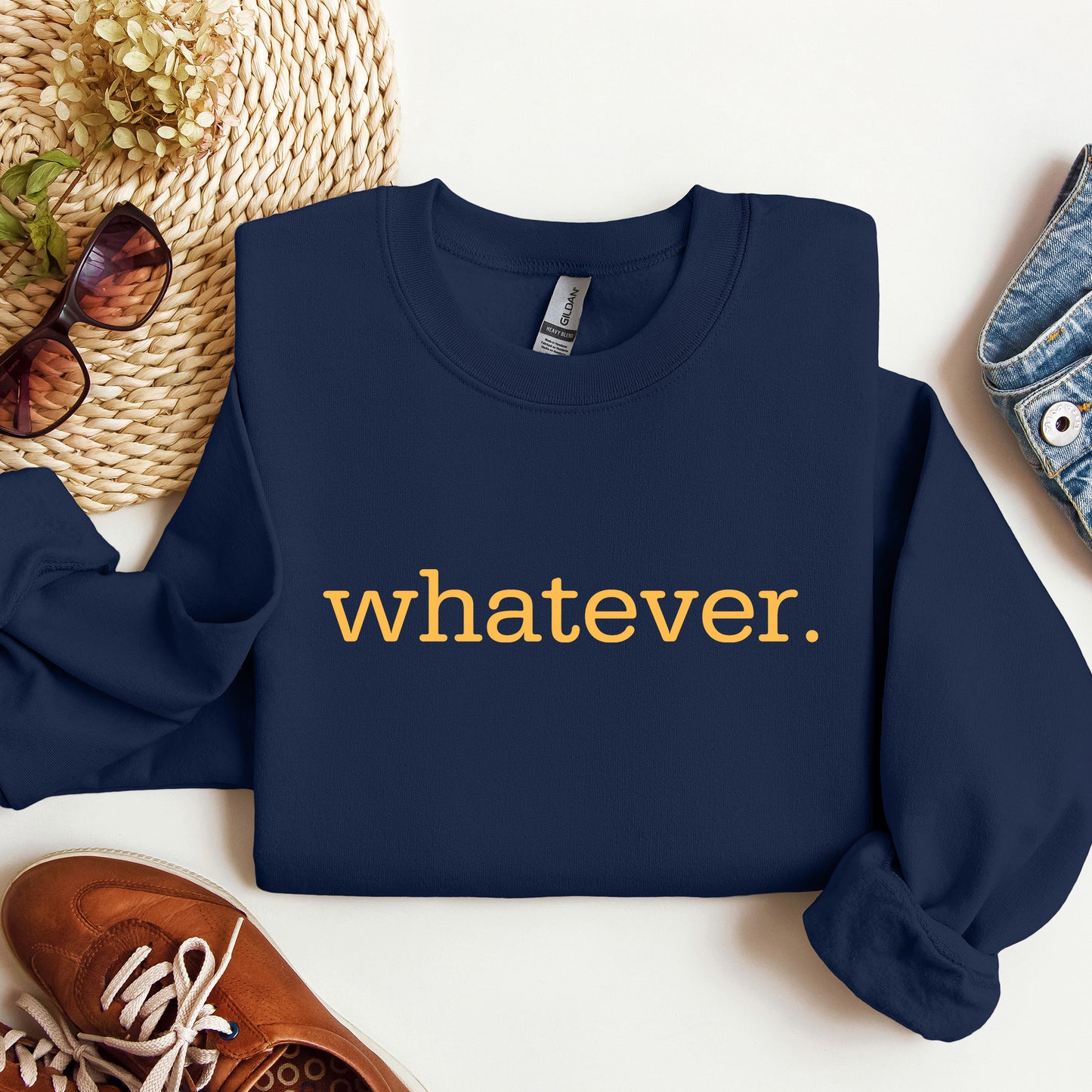 A navy blue crewneck sweatshirt with the word 'whatever.' printed in yellow lowercase text across the chest, styled in a flat lay with sunglasses, a woven hat, a brown shoe, and denim on a white background.