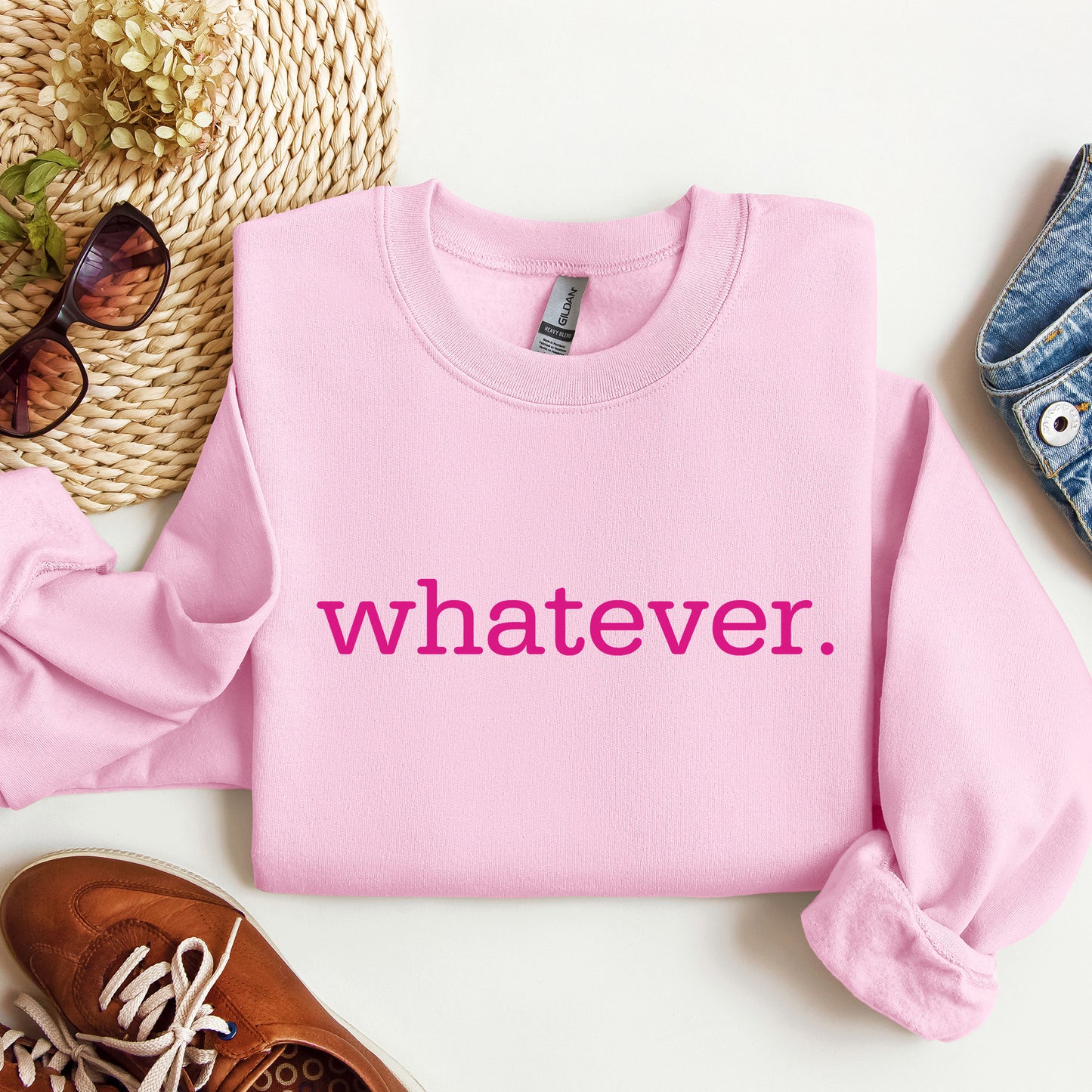 A folded light pink crewneck sweatshirt with the phrase 'whatever' printed in bright pink across the chest, styled in a flat lay with sunglasses, a woven hat, a brown shoe, and a denim item on a white background.