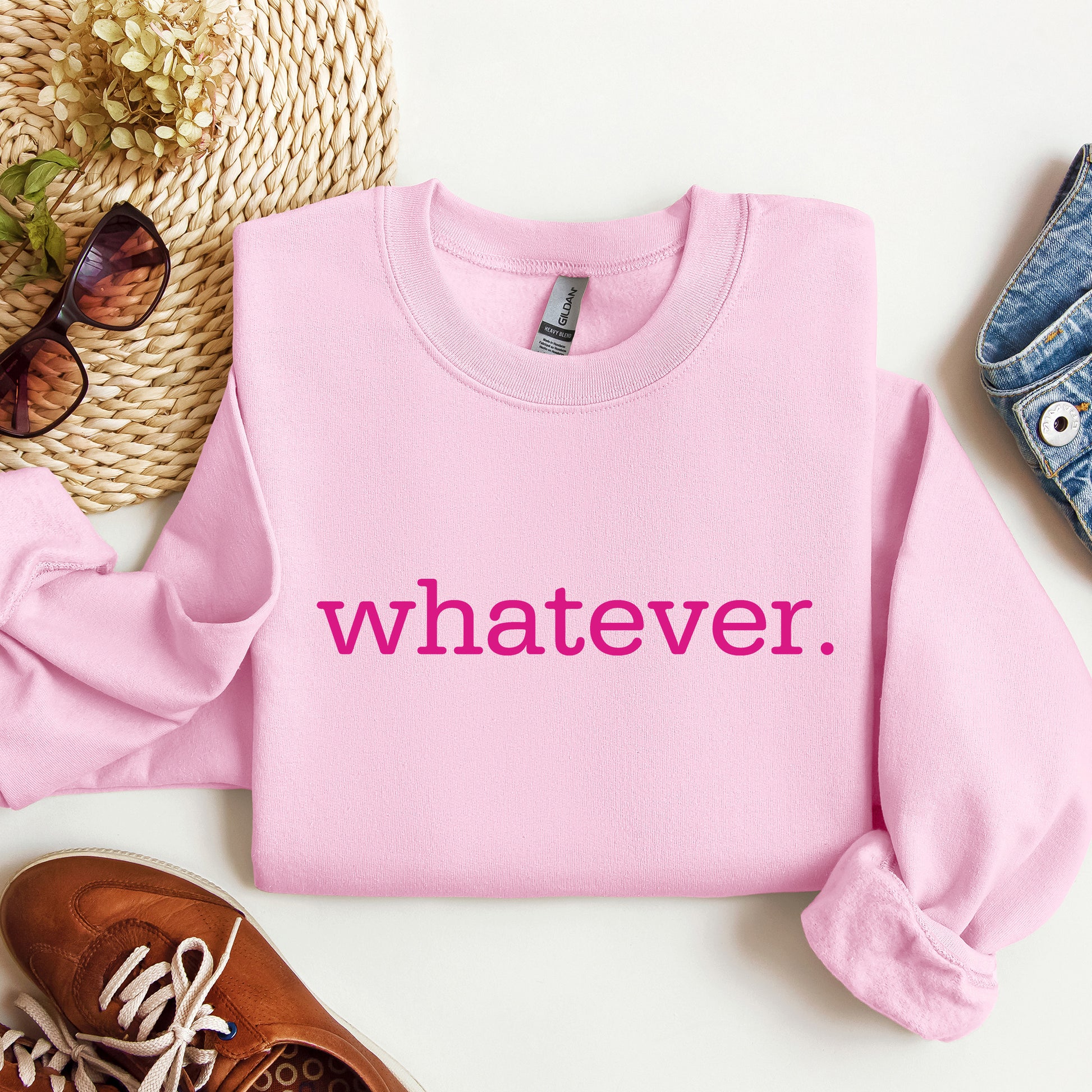 A folded light pink crewneck sweatshirt with the phrase 'whatever' printed in bright pink across the chest, styled in a flat lay with sunglasses, a woven hat, a brown shoe, and a denim item on a white background.