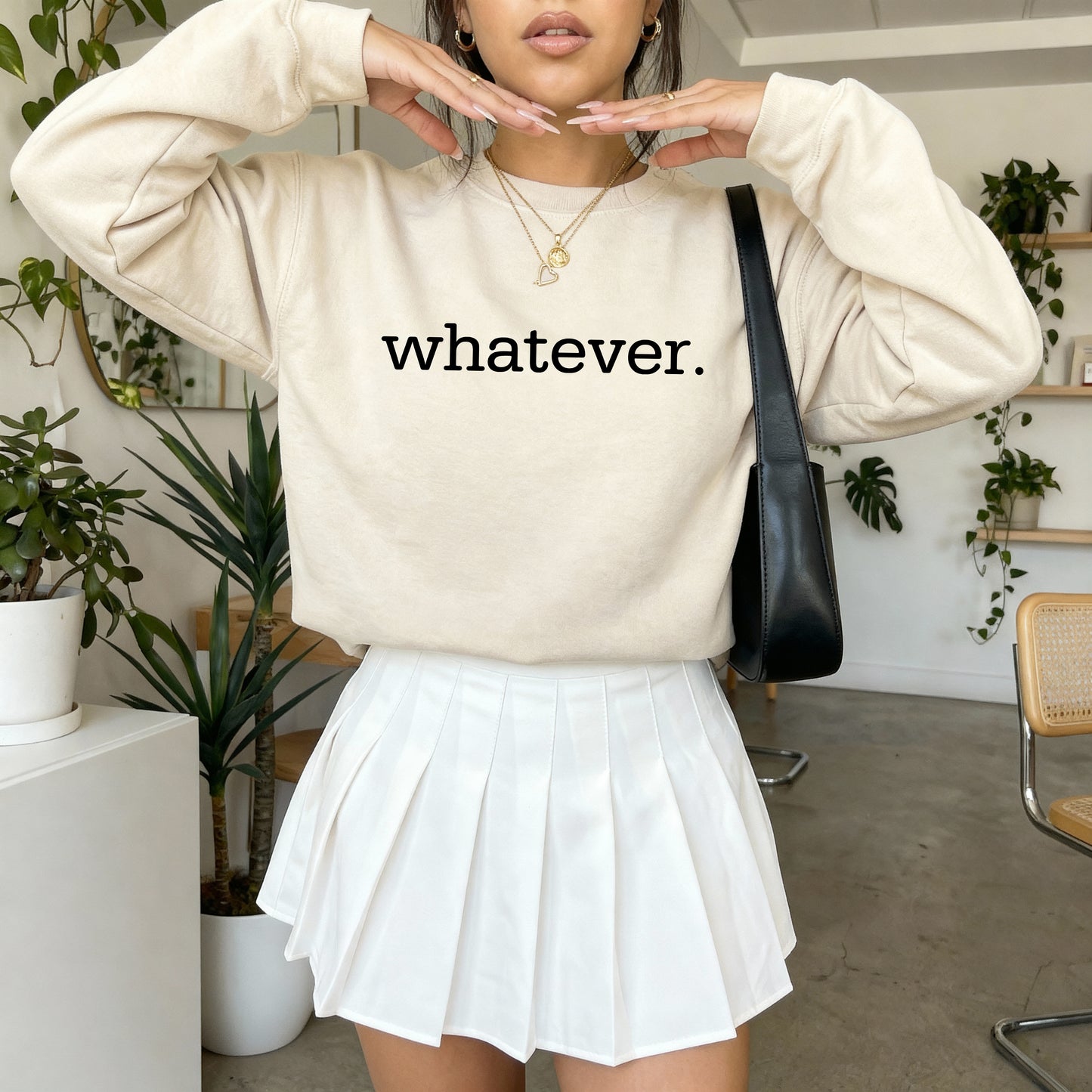 A woman wearing a cream-colored sweatshirt with the word 'whatever.' printed in black lowercase letters across the chest, paired with a white pleated mini skirt and a black shoulder bag, set in a bright indoor room with plants.
