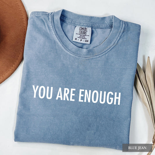 A folded light blue t-shirt with the phrase 'YOU ARE ENOUGH' printed in bold white capital letters across the chest. The t-shirt is displayed on a white surface with a brown hat on the left and dried leaves on the right.
