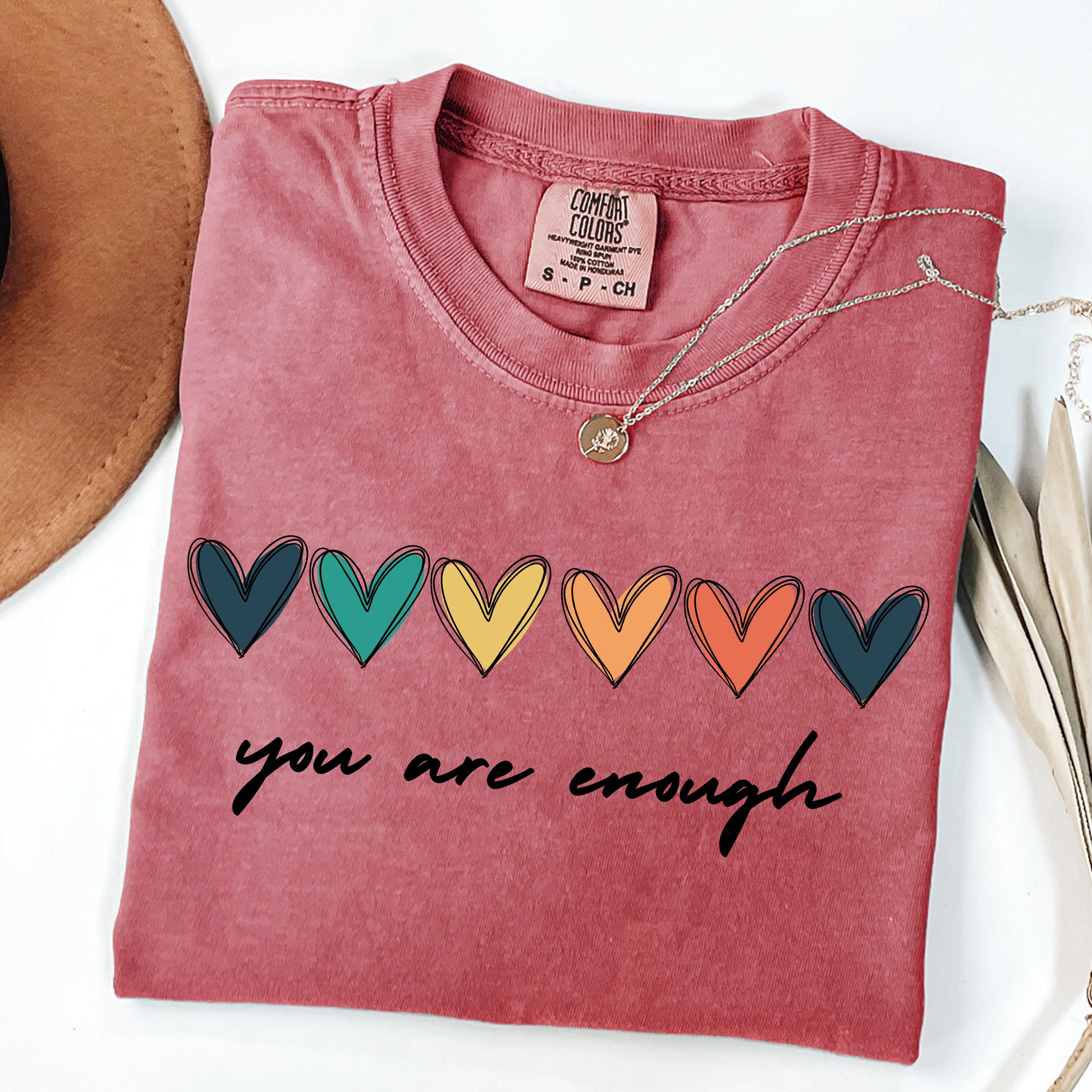 Folded pink t-shirt with colorful heart design and 'you are enough' text on a white background.