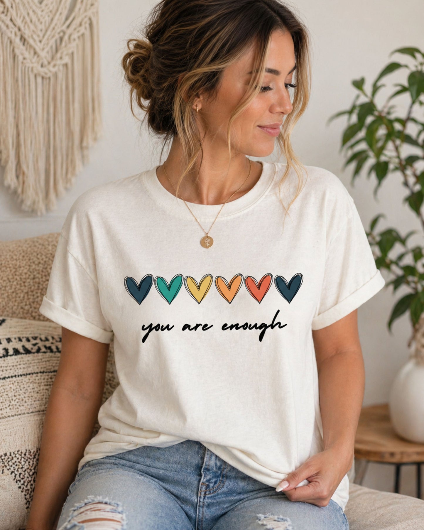 Woman wearing a white t-shirt with colorful heart designs and you are enough, sitting in a cozy room.