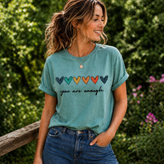 Woman wearing a teal t-shirt with colorful heart symbols and 'you are enough' text, standing outdoors.