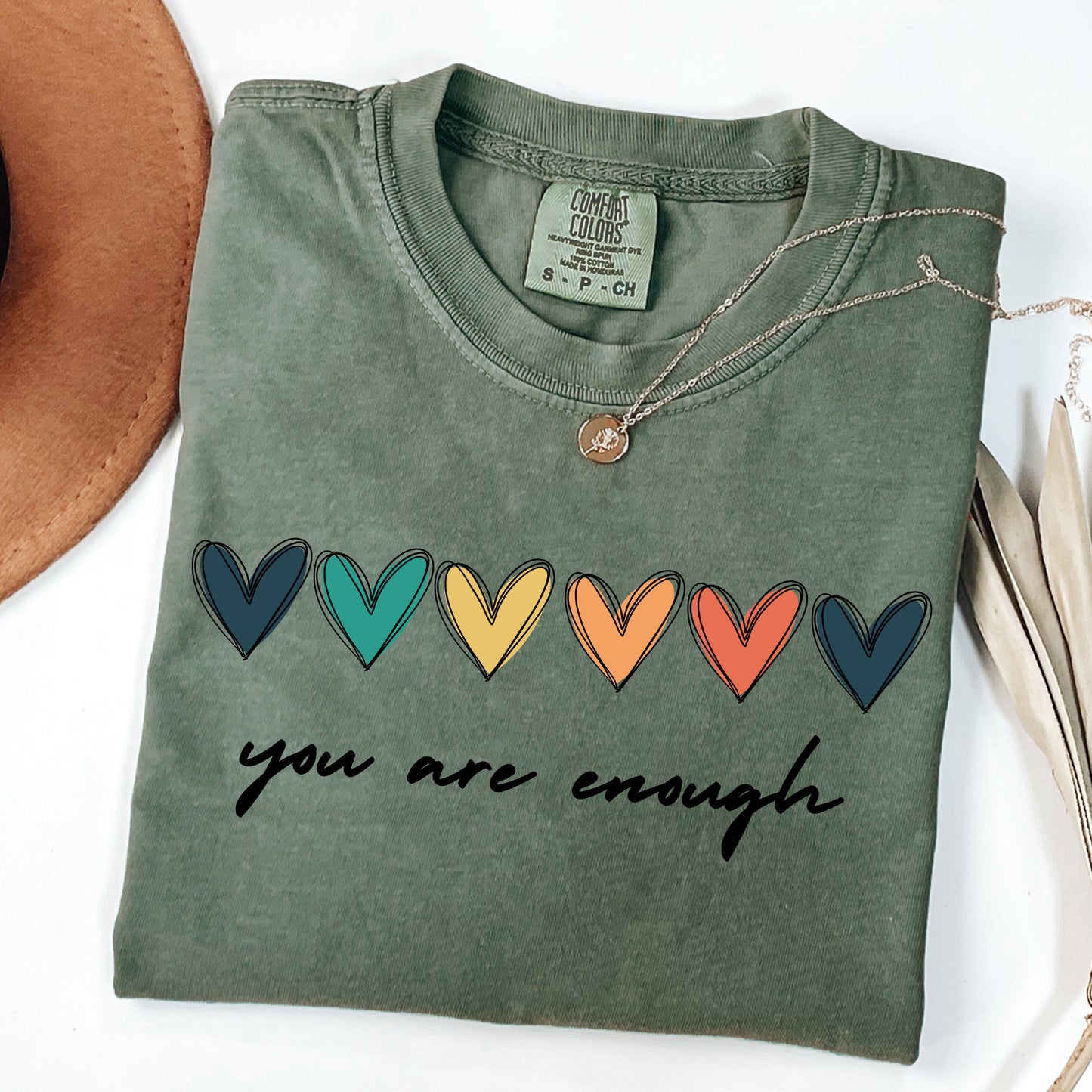 Folded green shirt with colorful hearts and 'you are enough' text on a white background