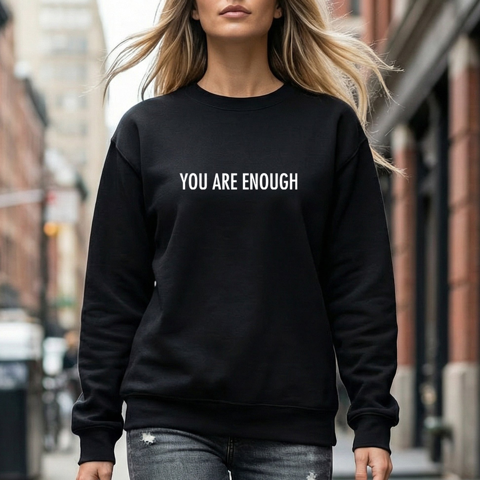 A woman wearing a black crewneck sweatshirt with the text 'YOU ARE ENOUGH' printed in white across the chest, standing on an urban street.