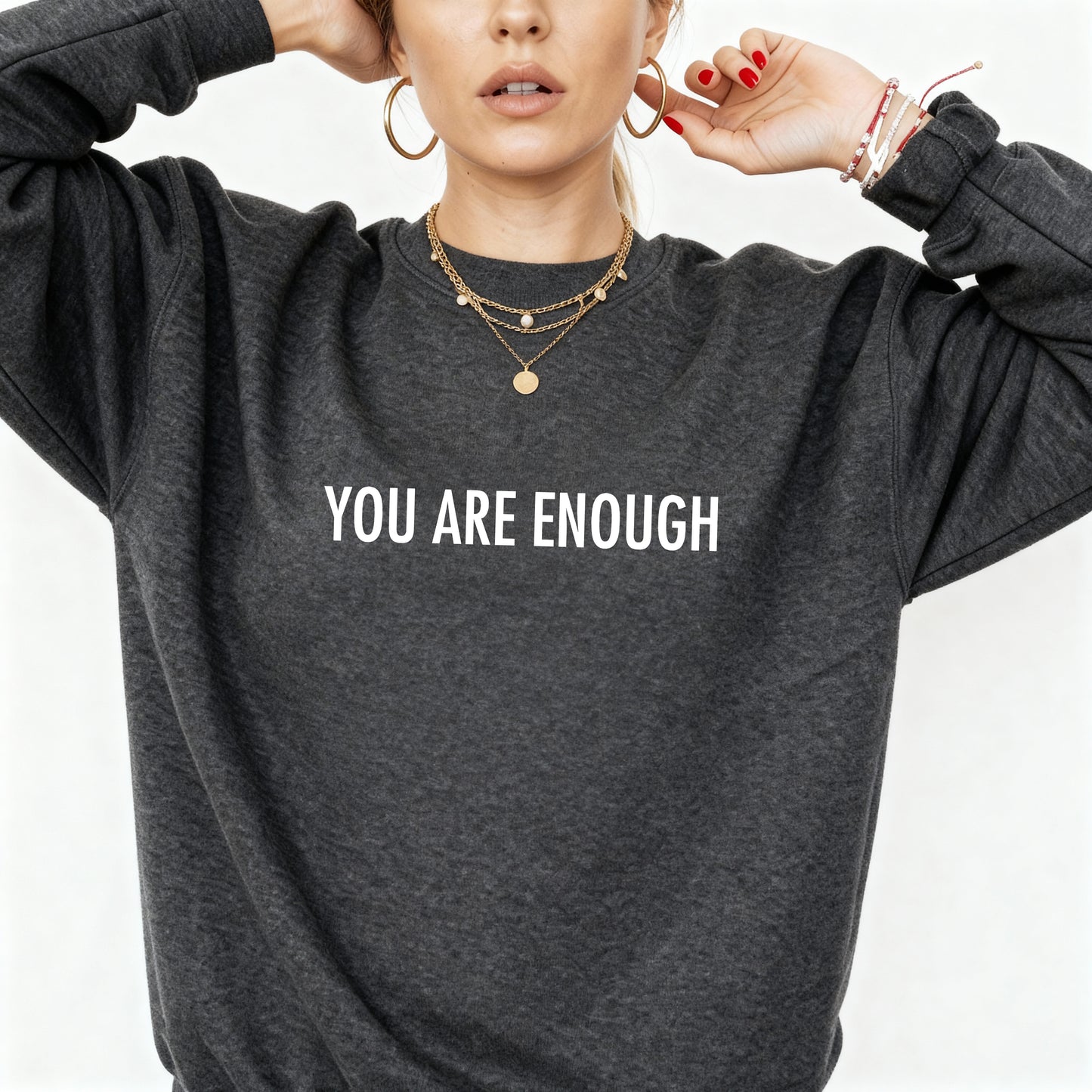 A woman wearing a dark heather gray crewneck sweatshirt with the words 'You Are Enough' printed in white serif text across the chest. She is wearing layered gold necklaces, hoop earrings, and bracelets against a clean white background.